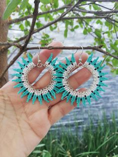 a person holding up two pairs of turquoise beaded hoop earrings in front of a tree