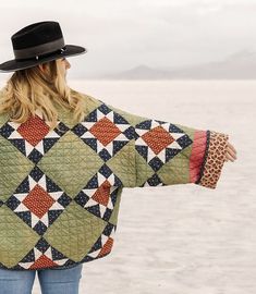 a woman wearing a quilted jacket and hat stands on the beach with her arms outstretched