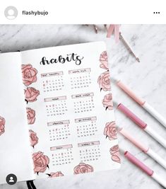 an open planner with pink flowers on it next to some pens and pencils in the background