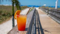 an orange drink sitting on top of a wooden table next to the beach and boardwalk