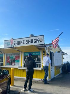 two men standing in front of a yellow shack with an american flag on the roof