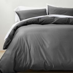 a bed with grey sheets and pillows on top of wooden floor next to white wall