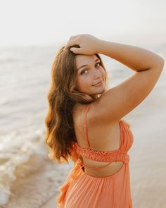 a woman in an orange dress standing on the beach with her arms behind her head