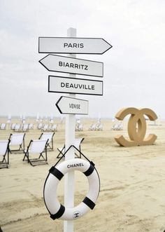 a sign on the beach with chairs in the background and a life preserver hanging from it's pole