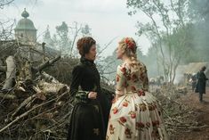 two women dressed in period clothing standing next to each other on a dirt road surrounded by trees