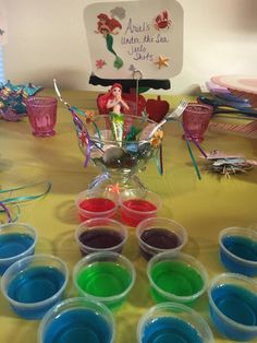 the table is covered with plastic cups filled with different colored liquids and little mermaid figurines