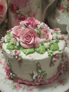 a white cake decorated with pink roses and green leaves on a plate in front of flowers