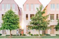 two people are walking down the sidewalk in front of some pink buildings with trees and bushes