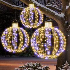 three christmas balls hanging from a tree in the snow with lights all around them on it