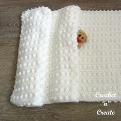 a teddy bear peeks out from behind a crocheted blanket on a wooden floor