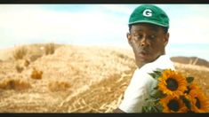 a man wearing a green hat and holding sunflowers in front of a field