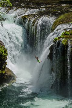 a person jumping off the side of a waterfall into a body of water with green moss growing on it