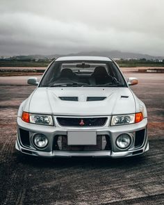 the front end of a white sports car parked in a parking lot with dark clouds overhead