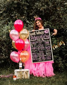 a woman standing next to a chalkboard sign with balloons on it in front of trees