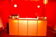 a bar with candles and bottles on it in front of a red wall that is lit up