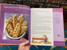 a hand holding an open book in front of a table full of food and bookshelves