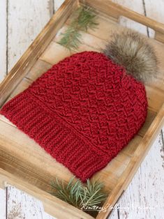 a red knitted hat with a pom - pom sits in a wooden box