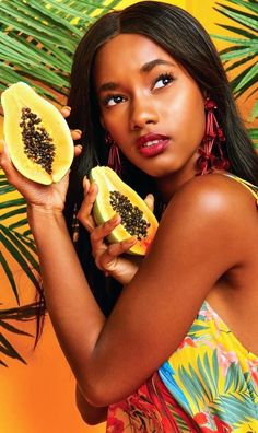 a beautiful young woman holding two pieces of fruit in front of her face and palm leaves behind her