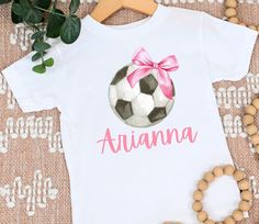 "Get ready to score big style points with this charming and personalized girls' soccer shirt! Your little soccer fan will look adorable and sporty with a soccer featuring a cute bow and her own custom name printed underneath. Customization: Make this shirt uniquely hers by adding her name! Simply leave the desired name in the \"Note to Seller\" section during checkout, and we'll create a one-of-a-kind shirt that she'll love to wear all season long. Quality and Comfort: Our shirts are crafted with the utmost care from soft and breathable cotton fabric, ensuring your little one's comfort during all her soccer-related activities. Whether she's cheering on her favorite team or playing a friendly game with friends, she'll feel at ease in this cozy and stylish shirt. Design Details: The watercol Personalized White Sporty T-shirt, Sporty Personalized T-shirt For Sports, Personalized Sporty T-shirt For Sports, Soccer Bow, Soccer Season, Soccer Tees, Soccer Outfit, Soccer Fan, Soccer Shirt