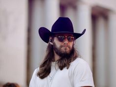 a man with long hair wearing a black cowboy hat and sunglasses standing in front of columns