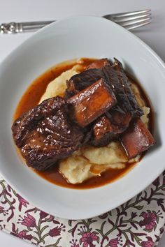 a white plate topped with meat and potatoes covered in gravy next to silverware