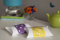 two pillows on a table next to a vase with flowers and teapot in the background