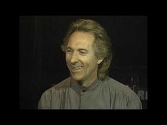 a man with long hair smiling at the camera in front of a black background and wearing a gray shirt