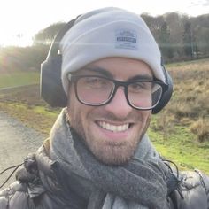 a man wearing glasses and a beanie smiles at the camera with headphones on