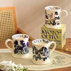 two coffee mugs sitting on top of a table next to a box and flowers