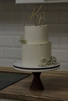 a three tiered cake with white flowers on top sits on a wooden table in front of a brick wall