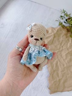 a hand holding a small crocheted teddy bear with a blue dress on it