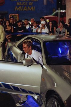 a man driving a silver sports car in front of a group of people on the street