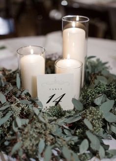 two candles sitting on top of a table covered in greenery and leaves with a place card next to it