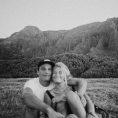 a man and woman sitting next to each other in the grass with mountains behind them