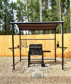 an outdoor bbq grill in the middle of a gravel area next to a wooden fence