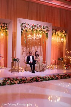 two people sitting on a couch in front of a stage with flowers and chandeliers