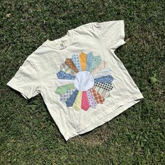 a white t - shirt with patchwork on it laying in the grass next to a pair of scissors