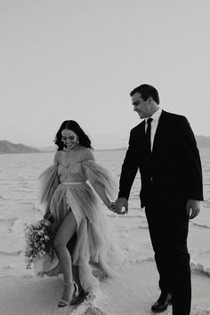 a man and woman walking on the beach holding hands, dressed in evening gowns