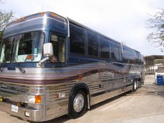 a large silver bus parked on top of a cement parking lot next to trees and bushes