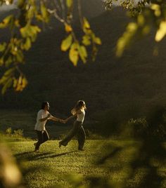 two people holding hands while running through the grass on a sunny day with trees in the background