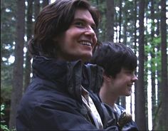 two men standing next to each other in the woods