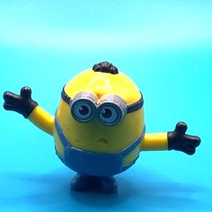 a yellow toy with black eyes and arms out on a blue surface, in the shape of a minion