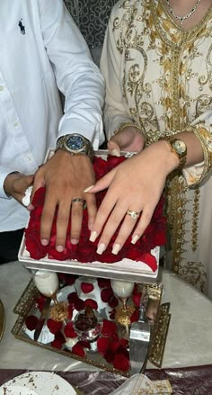 two people are cutting into a cake on a table with red flowers and gold trimmings