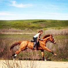 a woman riding on the back of a brown horse across a field with tall grass