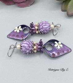 two purple earrings with flowers and pearls hanging from hooks on a table next to a flower