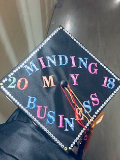 a black graduation cap that says minding my business on the front, and colorful writing on the back