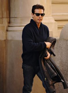 a man in black jacket and sunglasses walking down the street with his coat over his shoulder