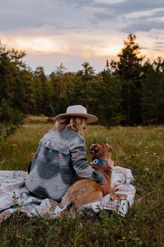 a woman sitting on top of a blanket with a dog next to her in the grass