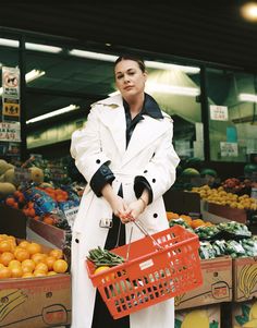 a woman in a white coat holding a red shopping basket next to crates of oranges