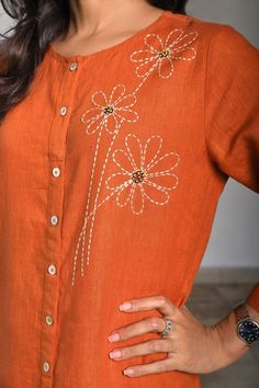 a woman wearing an orange shirt with white flowers on the front and side, holding her hands in her pockets